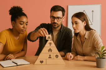 Três profissionais constroem uma pirâmide de madeira que simboliza a hierarquia de necessidades de Maslow sobre uma mesa. Ao fundo, quadro com desenho simples de pirâmide sem texto.