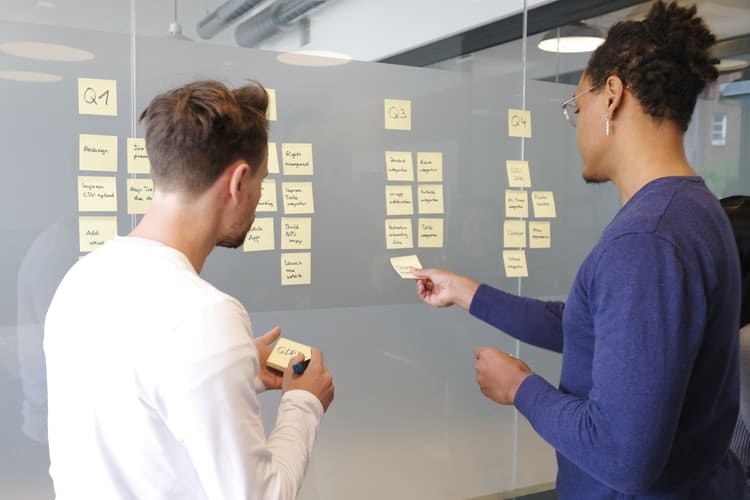 Imagem com dois profissionais analisando anotações de publicidade em post-its em um quadro. Faça uma análise da marca para a campanha de publicidade